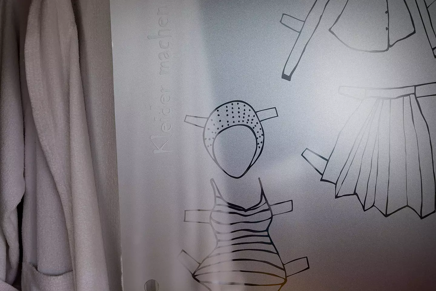 Detail. Auf einer Schrankschiebetür befindet sich eine Grafik von Kleidungstücken: Badeanzug, Badekappe, Faltenrock. Im linken Teil des Bildes sieht man Details von echte Kleidungsstücken aus dem Schrank.